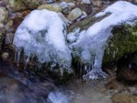 Steine am Bachufer bedeckt mit Eiskappe und Eisfiguren über dem Wasser
